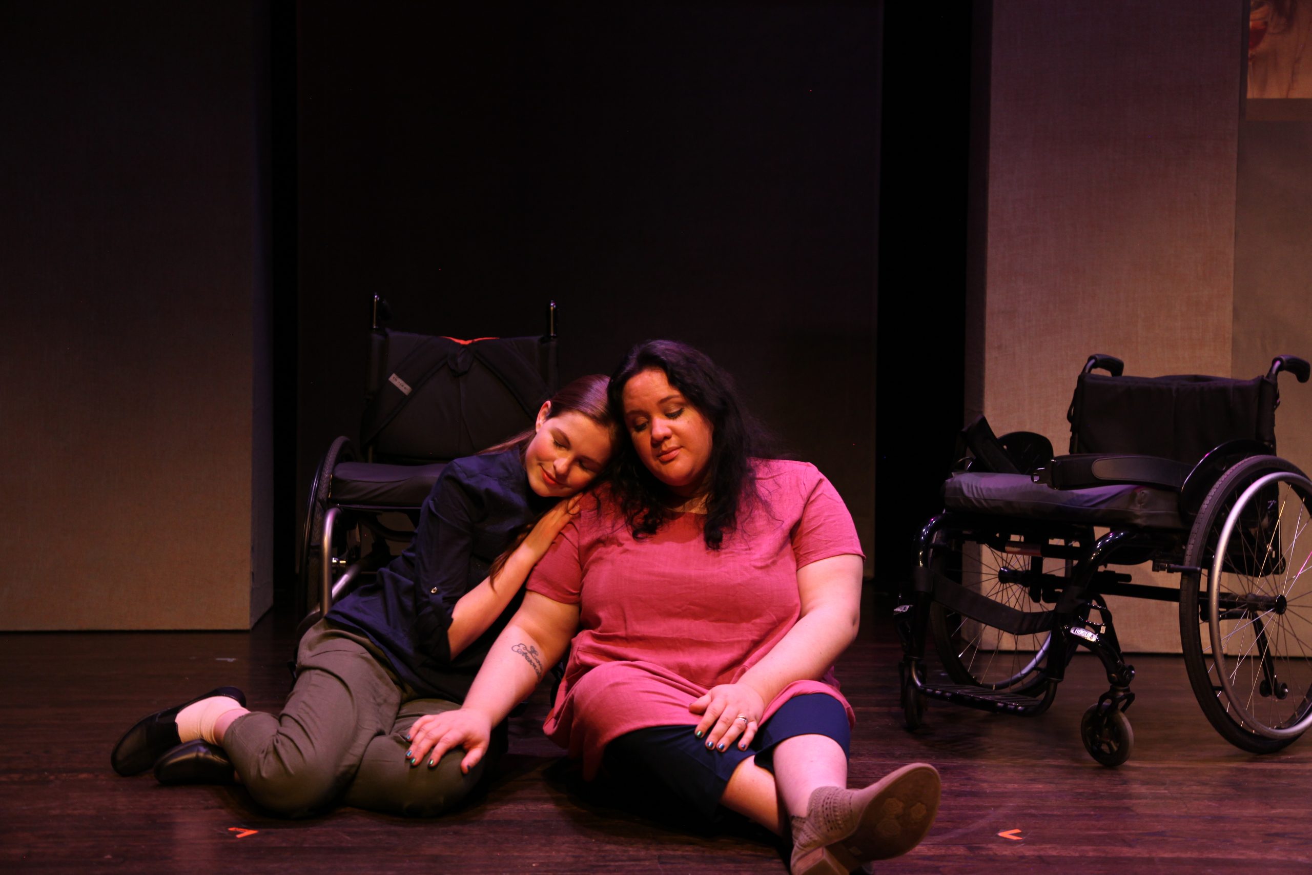 Production Photo from Act of Faith - Two actors sitting on the ground cuddling, their wheelchairs behind them.