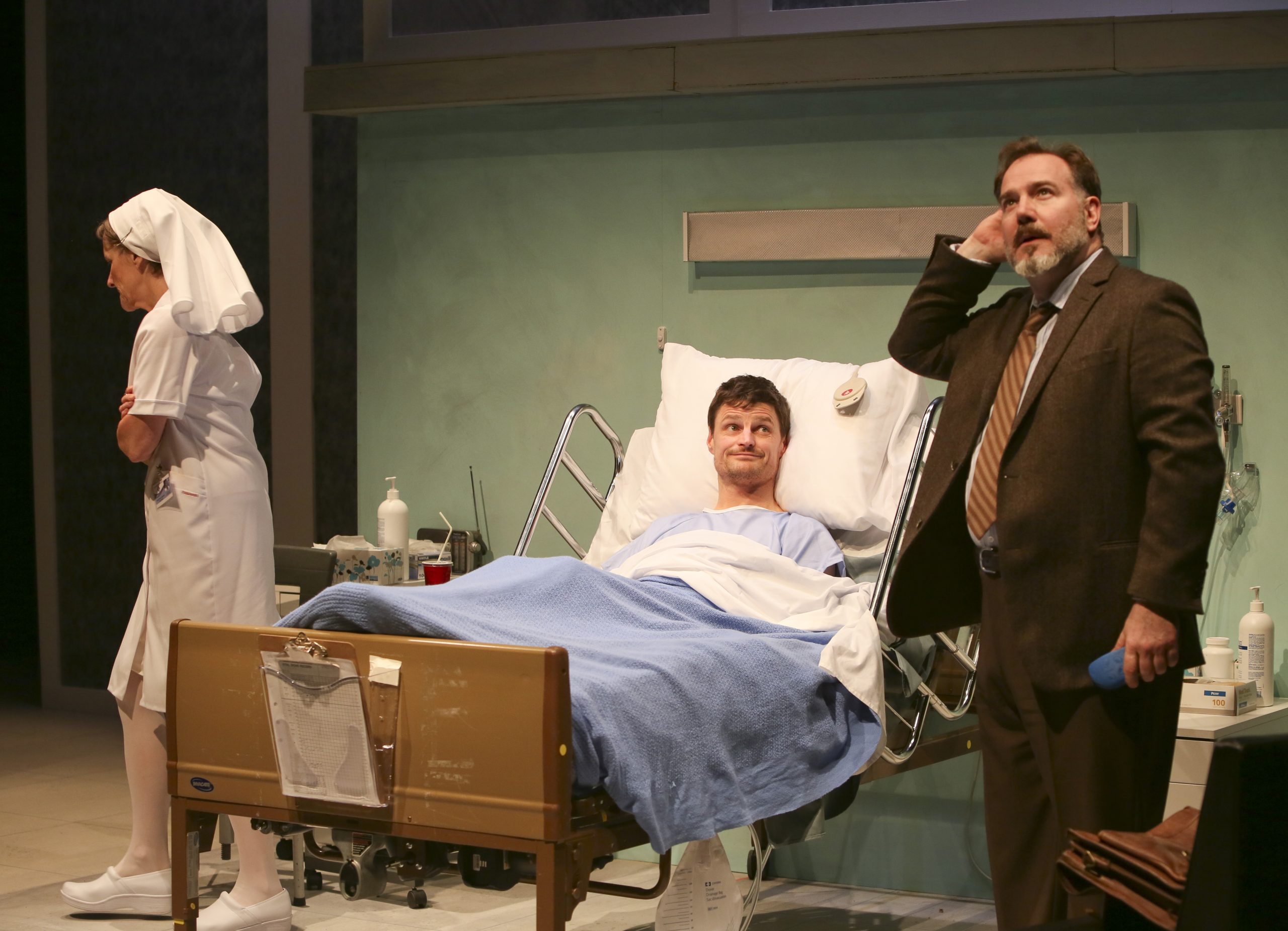 Production Photo from Whose Life is it Anyway? — Three actors, one in a hospital bed, the others a man in a suit and a nurse.