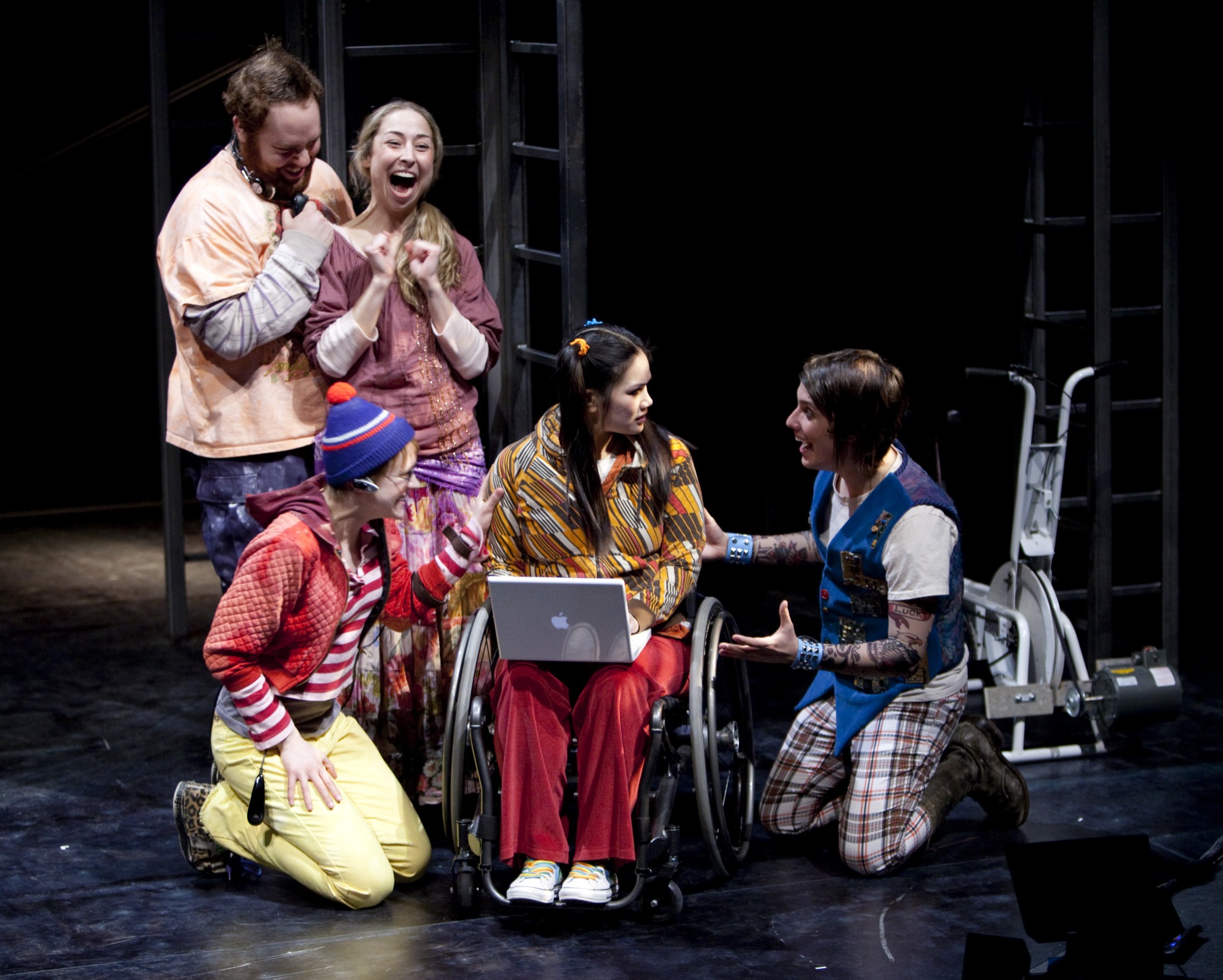 Production Photo from Spine - Five actors onstage, four looking excited, the fifth in a wheelchair with a laptop.