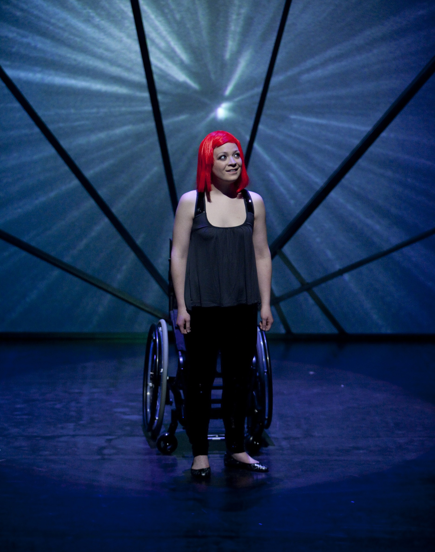 Production Photo from Spine - An actor in a red wig standing in front of a wheelchair