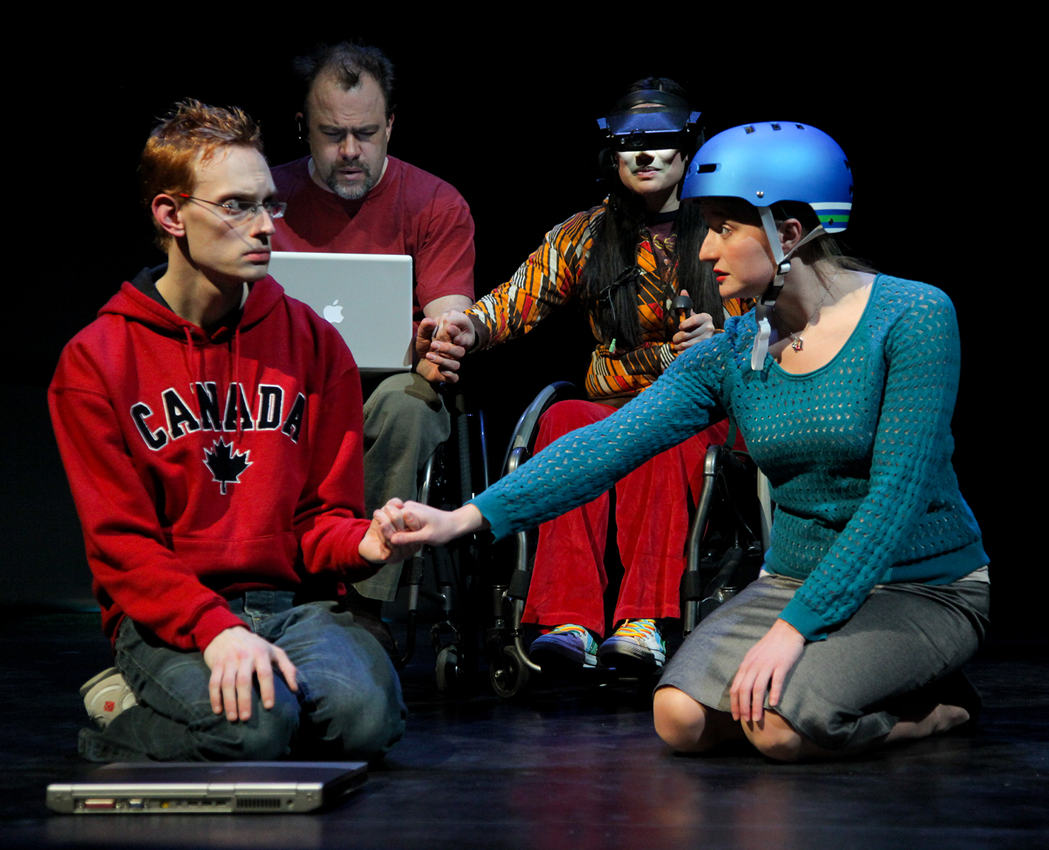 Production Photo from Spine - Four actors, one with a laptop, one with a VR headset, one with a bike helmet. They hold hands in pairs.