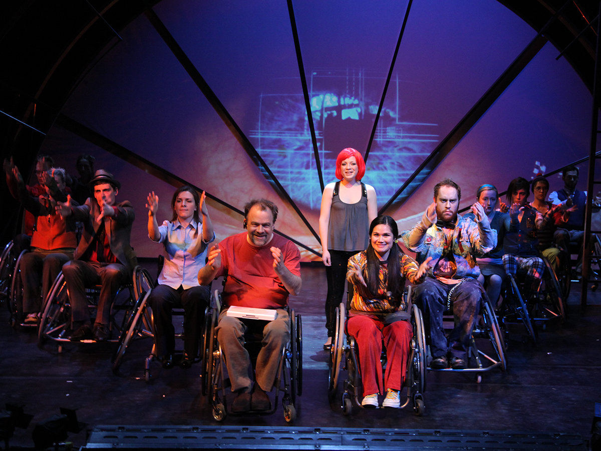 Production Photo from Spine - Full cast on stage, all except one in wheelchairs.