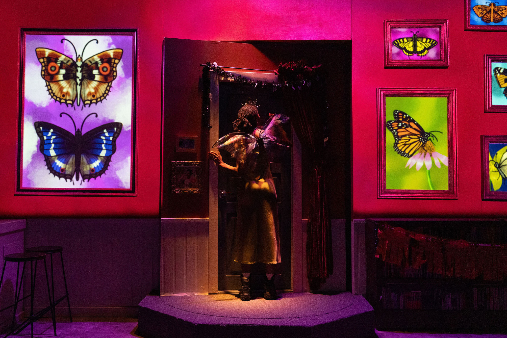 Production photo of Faye's Room - An actor going through a revolving door, framed butterflies on the wall.